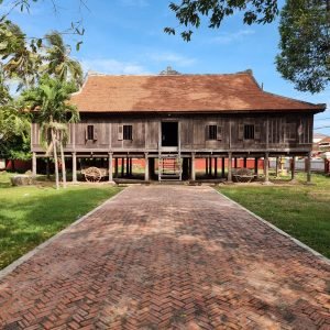 An old wooden Rokakondal Pagoda with a yellow exterior and traditional red-tiled roof, located in Kratie Town, Cambodia. The foreground features a weathered stone stupa, a brick-paved courtyard, and lush green trees under a partly cloudy blue sky.