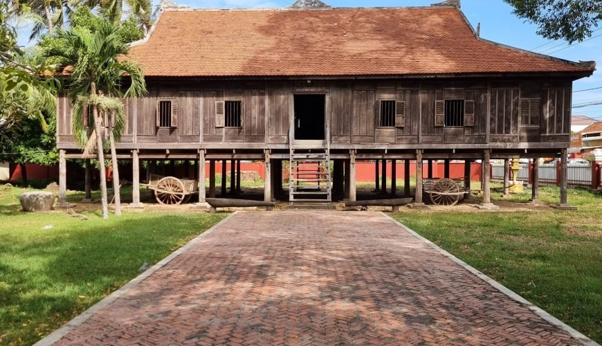 An old wooden Rokakondal Pagoda with a yellow exterior and traditional red-tiled roof, located in Kratie Town, Cambodia. The foreground features a weathered stone stupa, a brick-paved courtyard, and lush green trees under a partly cloudy blue sky.