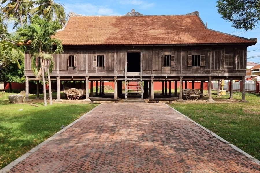 An old wooden Rokakondal Pagoda with a yellow exterior and traditional red-tiled roof, located in Kratie Town, Cambodia. The foreground features a weathered stone stupa, a brick-paved courtyard, and lush green trees under a partly cloudy blue sky.